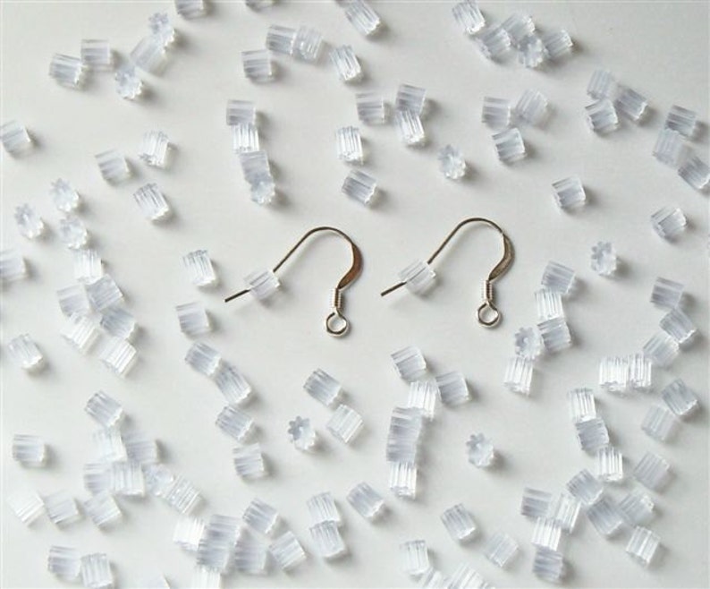 May include: A close-up of a white surface with a large quantity of clear plastic earring backs. Two silver earring hooks are also visible.