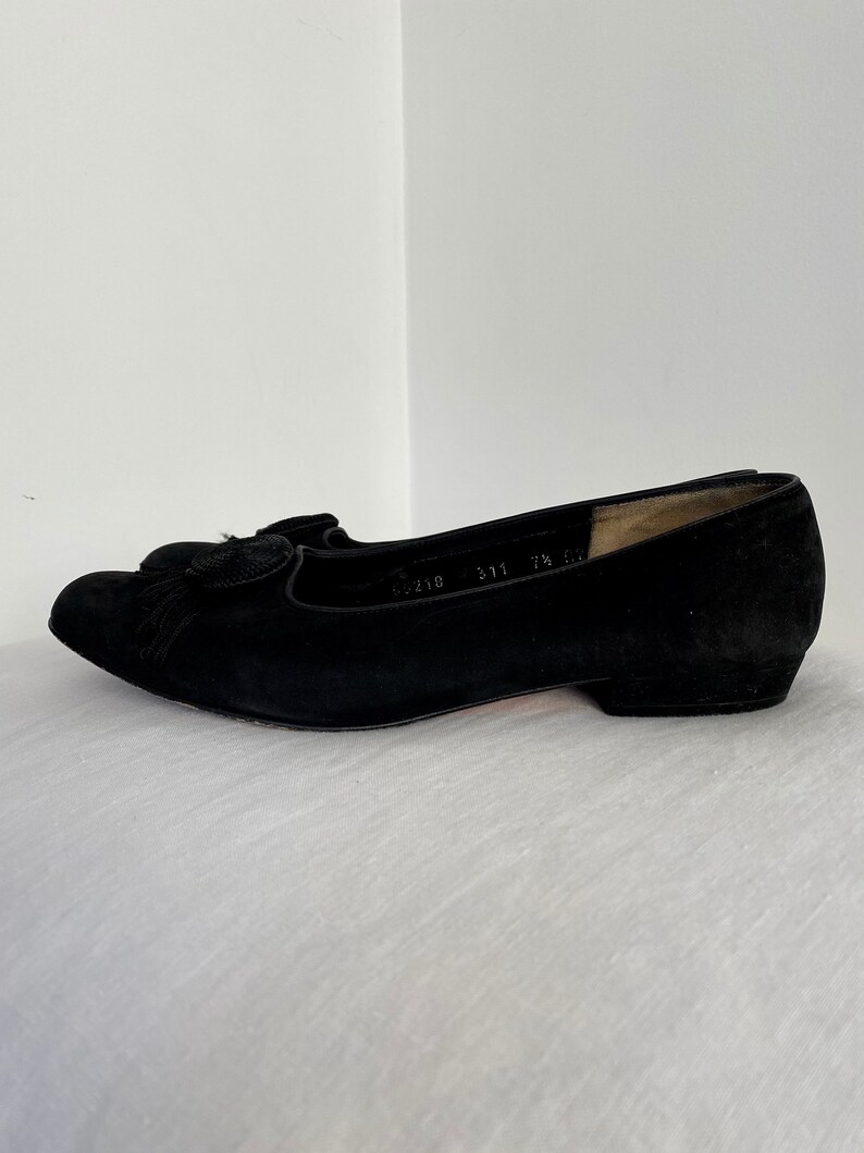 K&ouml;nnte beinhalten: Schwarze Ballerinas aus Wildleder mit einem kleinen Quasten-Detail an der Spitze. Die Ballerinas haben einen niedrigen Absatz und sind mit der Gr&ouml;&szlig;e 311 7 1/2 gekennzeichnet.