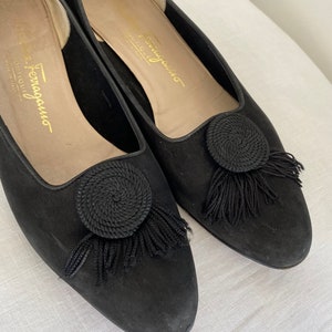 K&ouml;nnte beinhalten: Ein Paar schwarze Wildlederschuhe mit einem kleinen schwarzen Quasten und einem kreisf&ouml;rmigen Design an der Spitze. Die Schuhe haben eine goldene Inschrift mit der Aufschrift "Salvatore Ferragamo" auf der Innenseite.