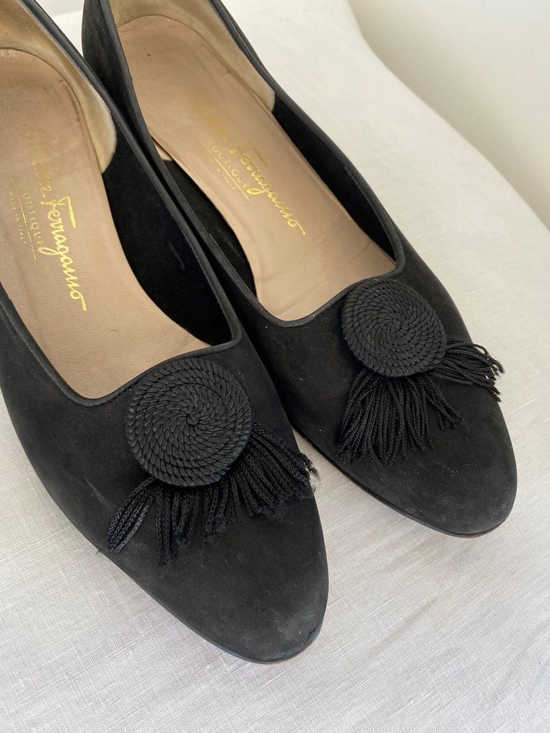 K&ouml;nnte beinhalten: Ein Paar schwarze Wildlederschuhe mit einem kleinen schwarzen Quasten und einem kreisf&ouml;rmigen Design an der Spitze. Die Schuhe haben eine goldene Inschrift mit der Aufschrift "Salvatore Ferragamo" auf der Innenseite.