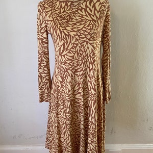 Könnte beinhalten: Ein langärmeliges Kleid mit einem abstrakten Blatt-Muster in Braun und Creme. Das Kleid hat einen runden Ausschnitt und einen fließenden Rock.