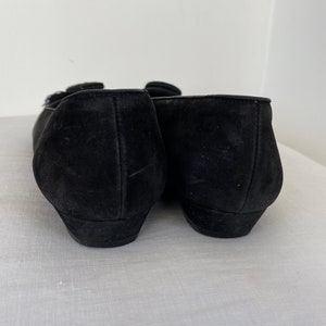 K&ouml;nnte beinhalten: Ein Paar schwarze Ballerinas aus Wildleder mit einem kleinen Schnallen-Detail an der Spitze.