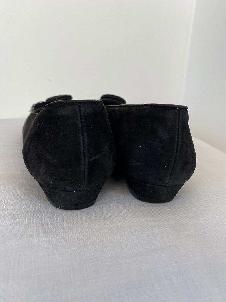K&ouml;nnte beinhalten: Ein Paar schwarze Ballerinas aus Wildleder mit einem kleinen Schnallen-Detail an der Spitze.