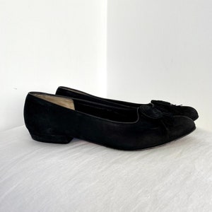K&ouml;nnte beinhalten: Ein Paar schwarze Ballerinas aus Wildleder mit einem kleinen schwarzen Quasten-Detail an der Spitze.