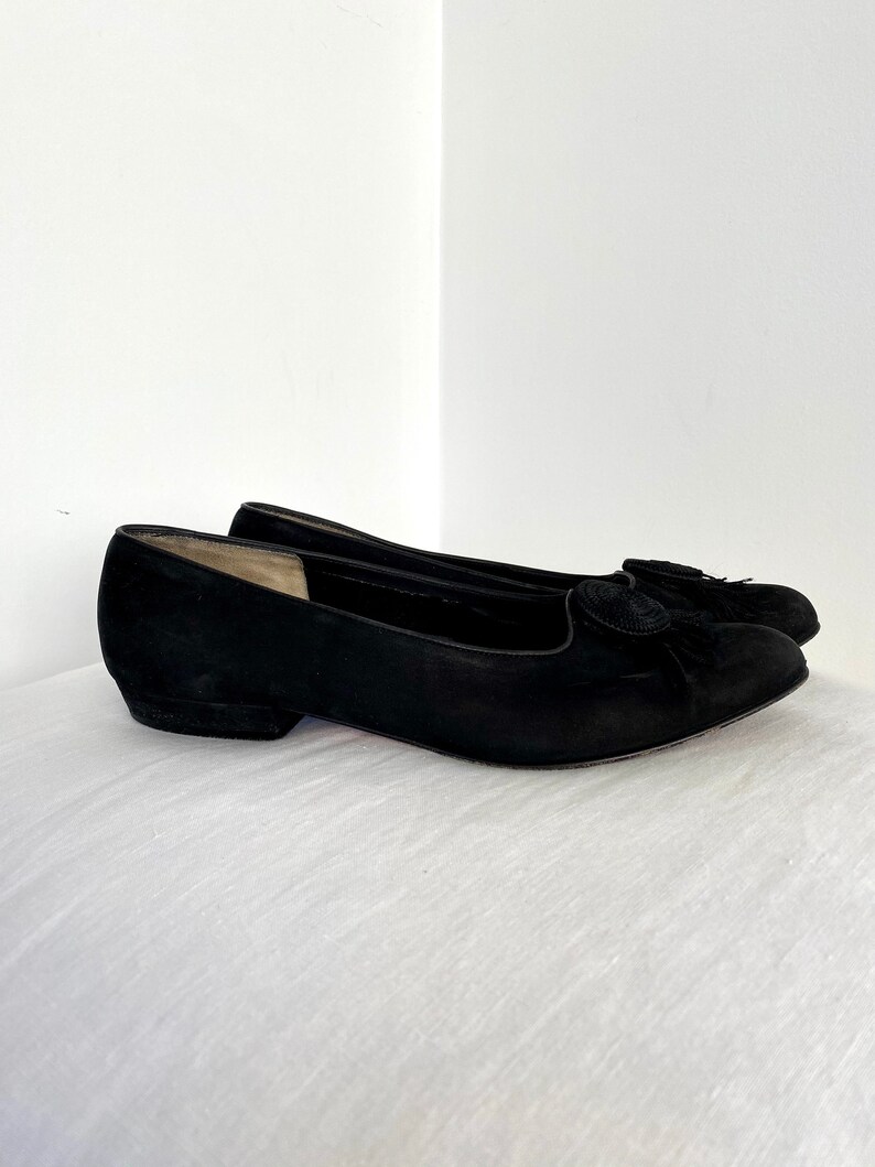 K&ouml;nnte beinhalten: Ein Paar schwarze Ballerinas aus Wildleder mit einem kleinen schwarzen Quasten-Detail an der Spitze.