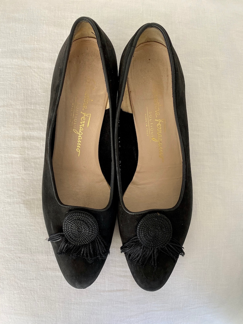 K&ouml;nnte beinhalten: Ein Paar schwarze Wildlederschuhe mit einem schwarzen Quasten und einem schwarzen Seildetail an der Spitze. Die Schuhe sind innen mit "Salvatore Ferragamo" beschriftet.