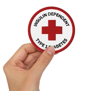 Op de afbeelding: Een ronde patch met een rode rand en een witte binnenkant. De patch toont een rood kruis en de woorden "INSULIN DEPENDENT" en "TYPE 1 DIABETES" in zwarte tekst. De patch wordt in een hand gehouden.