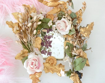 Op de afbeelding: Een vintage bloemstuk met een kleine spiegel in het midden. Het arrangement bevat roze rozen, witte bloemen, gouden bladeren en groen blad.