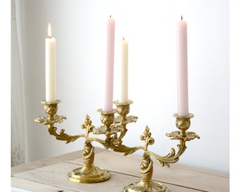 Op de afbeelding: Een paar gouden metalen kandelaars met sierlijke ontwerpen. Elke kandelaar heeft drie kaarsarmen en is versierd met ingewikkelde bloemmotieven. De kaarsen zijn roze en wit.