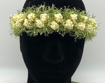 May include: A floral crown featuring off-white, clustered flower buds set against a backdrop of green, textured foliage. The crown is displayed on a black mannequin head, suggesting a bridal or special occasion accessory.