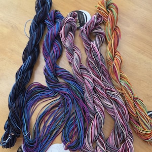 May include: Five hanks of multi-colored embroidery thread arranged side by side. The threads feature variegated color combinations including blue and black, purple and blue, pink and white, purple and white, and orange, yellow, and green tones.
