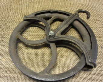 May include: A vintage, cast iron pulley wheel with a hook. The wheel has a circular design with spokes and a central bolt. The metal has a weathered, rustic appearance, with visible signs of age and use. The pulley is designed for rope or cable.