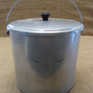 May include: A vintage aluminum bucket with a lid and a metal handle. The bucket is cylindrical with a silver finish and a black knob on the lid. The handle arches over the top. The bucket is likely used for carrying water or other items.