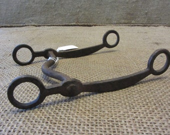 May include: A dark brown metal horse bit with a curved design. It has two circular rings at each end and a curved mouthpiece. The metal has a rustic, aged appearance, suggesting it is antique. The bit is likely for a horse.