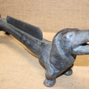 May include: A dark gray cast iron boot scraper shaped like a long-bodied dog. The object features a raised vertical blade along the center of the back, a curled tail, and a head with an open mouth, all cast in a textured, weathered metal finish.