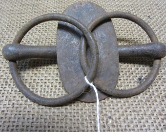 May include: A rusty metal horse bit with two rings. The bit is oval shaped and has a rounded bar connecting the rings.