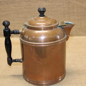 Peut inclure: Une théière cylindrique en cuivre avec une finition métallique polie, une poignée en bois noir foncé au design vertical et un bouton en bois noir assorti sur le couvercle. La théière comprend un bec court et large ainsi qu'un couvercle étagé.