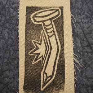 以下が含まれることがあります： 星形の背景にスタイリッシュな釘の画像が描かれた、茶色と黒の布のパッチです