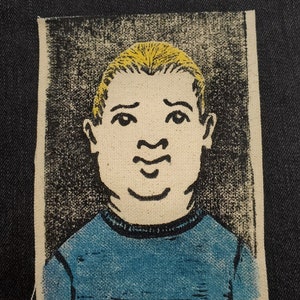 Pode incluir: Remendo de tecido retangular com uma ilustração estilizada de um rosto. A pessoa tem cabelo amarelo e uma camisa azul. O fundo é cinza texturizado e as bordas são bordadas de branco. A expressão facial é neutra.