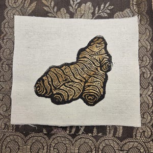 May include: A square piece of off-white fabric featuring a screen-printed illustration of a ginger root. The ginger root is rendered in shades of brown and tan with dark black outlines, showcasing a textured, organic, and knobby appearance.