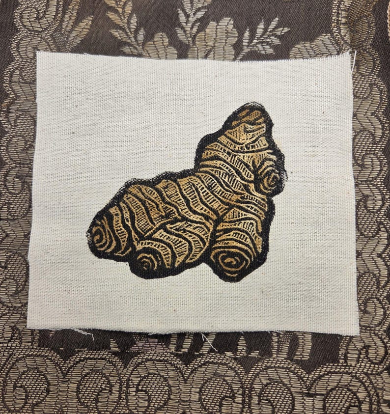 May include: A square piece of off-white fabric featuring a screen-printed illustration of a ginger root. The ginger root is rendered in shades of brown and tan with dark black outlines, showcasing a textured, organic, and knobby appearance.