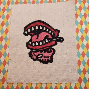 Könnte beinhalten: Ein pink-rotes Cartoon-Mund-Motiv auf einem Leinwand-Patch. Der Mund ist geöffnet und zeigt Zähne und ein pinkfarbenes Inneres. Der Patch befindet sich auf einem farbenfrohen Rautenmuster-Hintergrund.