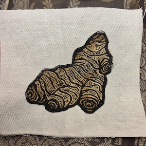 May include: A rectangular piece of off-white canvas fabric featuring a centered, detailed screen-printed illustration of a ginger root. The ginger root is rendered in shades of brown and tan with bold black outlines and textured, organic lines.