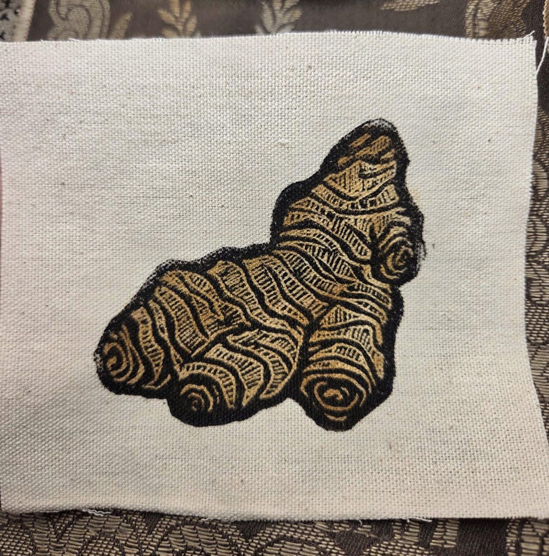 May include: A rectangular piece of off-white canvas fabric featuring a centered, detailed screen-printed illustration of a ginger root. The ginger root is rendered in shades of brown and tan with bold black outlines and textured, organic lines.