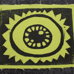 May include: A black and green fabric patch with a stylized sun design. The sun has a black center with a circle around it and a ring of points around the circle.