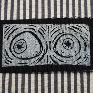 May include: Black and white embroidered patch with two large eyes. The eyes are surrounded by swirling lines and have black pupils.