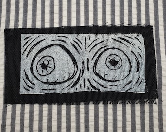May include: Black and white embroidered patch with two large eyes. The eyes are surrounded by swirling lines and have black pupils.