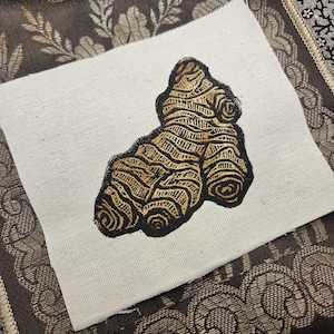 May include: A square piece of off-white canvas fabric featuring a block-printed illustration of a ginger root. The ginger root is rendered in a metallic gold ink with bold black outlines, showcasing the textured, segmented surface of the root.