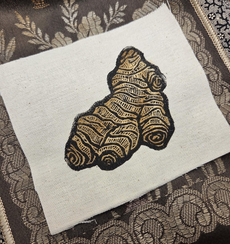 May include: A square piece of off-white canvas fabric featuring a block-printed illustration of a ginger root. The ginger root is rendered in a metallic gold ink with bold black outlines, showcasing the textured, segmented surface of the root.