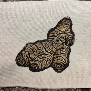 May include: A piece of off-white canvas fabric featuring a detailed, dark brown embroidered ginger root. The embroidery uses thick, bold lines to create a textured, stylized representation of the ginger, centered on the light-colored textile.