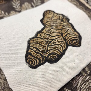 May include: A square piece of off-white canvas fabric featuring a black and gold block print illustration of a ginger root. The ginger root design displays intricate, textured line work and organic, knobby shapes centered on the fabric square.
