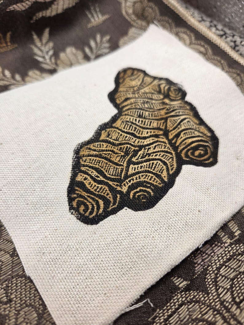 May include: A square piece of off-white canvas fabric featuring a black and gold block print illustration of a ginger root. The ginger root design displays intricate, textured line work and organic, knobby shapes centered on the fabric square.