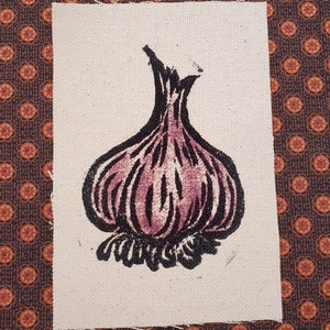 May include: A hand-printed image of a purple garlic bulb on a beige fabric background.