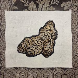 May include: A rectangular piece of off-white fabric featuring a centered, detailed illustration of a ginger root. The ginger root is rendered in shades of brown and gold with distinct, dark outlines that emphasize its knobby, textured surface.