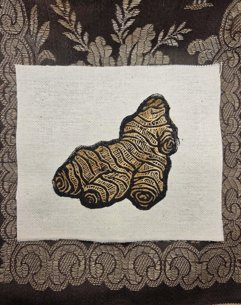 May include: A rectangular piece of off-white fabric featuring a centered, detailed illustration of a ginger root. The ginger root is rendered in shades of brown and gold with distinct, dark outlines that emphasize its knobby, textured surface.