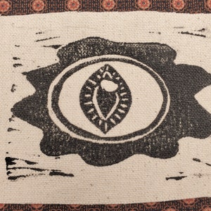 Puede incluir: Una impresión en blanco y negro de un ojo estilizado sobre un fondo de tela beige. El ojo está rodeado de un círculo y tiene una forma de hoja en su interior.