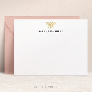 Könnte beinhalten: Weiße Briefpapierkarte mit einer goldenen Bienenillustration und dem Text "SARAH LIONSHead" in Schwarz gedruckt.
