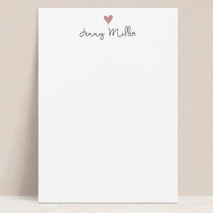 May include: A white notepad with a small pink heart above the handwritten name "Jenny Miller" in black script. The notepad is on a neutral background.