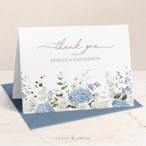 May include: A folded white card with the words "thank you" and "from REBECCA SANDERSON" in script. The bottom edge features a watercolor floral design with blue hydrangeas, white roses, and greenery. A blue envelope is visible.