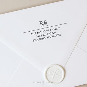 May include: White envelope with the address "THE MORGAN FAMILY, 1432 CURIO LN, ST. LOUIS, MO 63122" printed in black. A white wax seal with a bow design is in the lower right corner. The letter "M" is at the top.