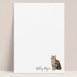May include: White notepad with a tabby cat illustration and the name "Ashley Harper" written in black cursive.