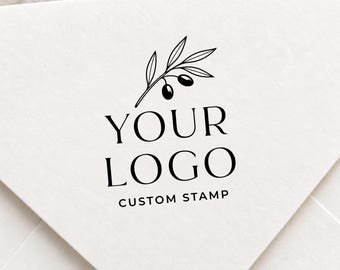 May include: A black ink stamp impression on white paper featuring a line art illustration of an olive branch with two olives. The text reads YOUR LOGO in large serif font, with the words CUSTOM STAMP printed in smaller sans-serif letters below.