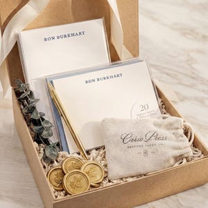 May include: A gift box with stationery, a pen, and wax seals. The box contains notecards and envelopes with the name "RON BURKHART" printed on them. A gold pen and wax seals with a leaf design are also included. A small bag with the text "Curio Press" is also in the box.