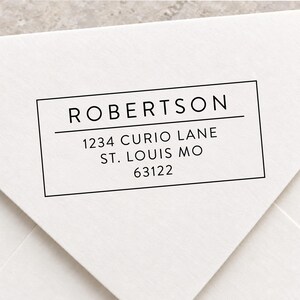 May include: A rectangular black ink stamp impression on a white envelope flap. The text reads ROBERTSON, followed by a horizontal line, then 1234 CURIO LANE, ST. LOUIS MO, and 63122. The design is enclosed within a thin black rectangular border.