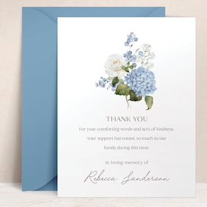 May include: A white card with a blue envelope. The card features a watercolor floral arrangement of blue hydrangeas, white roses, and greenery. Text on the card reads "THANK YOU" and a message of gratitude, with the name "Rebecca Sanderson".