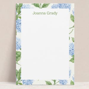 May include: Stationery with a floral design. The border features blue hydrangea blooms and green leaves on a cream background. The text "Joanna Grady" is printed in green at the top.